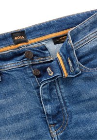 Smalnande blå jeans i denim med en orange detalj vid midjebandet, utrustade med två svarta knappar och en svart etikett märkt "BOSS".