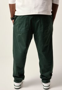 Groene katoenbroek met een relaxed fit, voorzien van twee achterzakken en een manchet onderaan, gecombineerd met witte en groene sneakers.