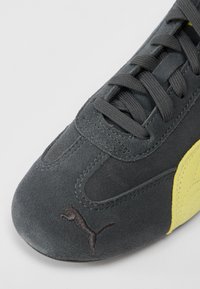 Lähedalt vaade hallile seemisnahast tossu varbale, millel on must Puma logo, tumehallad paelad ja kollane külgede paneeli detail.