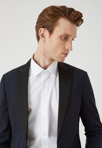 Giacca da smoking navy con rever in satin nero, abbinata a una camicia bianca con colletto strutturato e chiusura a bottoni.
