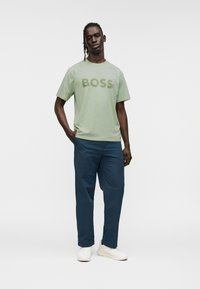 Ανοιχτό πράσινο κοντομάνικο μπλουζάκι με την επιγραφή "BOSS" σε πιο σκουρό πράσινο, συνδυασμένο με σκούρο μπλε παντελόνι και λευκά αθλητικά παπούτσια. Απλός σχεδιασμός, άνετη εφαρμογή.