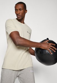 Beige kortærmet t-shirt med en broderet logo, parret med grå svedbukser, som holder en sort medicinbold. Glat tekstur, afslappet pasform.