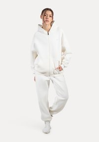 Hvid zip-hoodie med store lommer og hætte med snøre, parret med matchende sweatpants. Blød tekstur, oversized design, minimal branding.