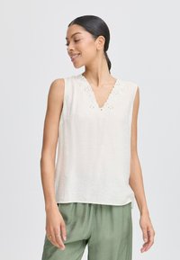 Top senza maniche di un leggero color crema con scollatura a V e bordo ricamato a onde. Tessuto morbido e fluido, abbinato a pantaloni verdi a vestibilità ampia.