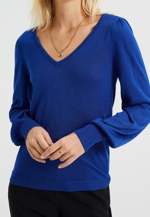Pull en tricot bleu avec un décolleté en V, des manches bouffantes et des poignets et un ourlet côtelés. Des fils pailletés ajoutent de la texture ; porté par-dessus un pantalon sombre.