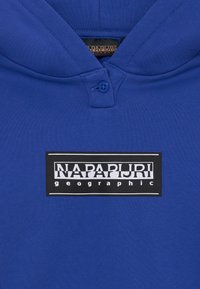 Blue polo shirt with a soft texture, featuring a black rectangular logo that reads "NAPAPURI geographic." Two-button placket at the collar.