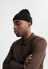 Zwarte breien beanie en een bruine langemouwen polo met een knoopkraag, met gekruiste armen en een neutrale achtergrond.