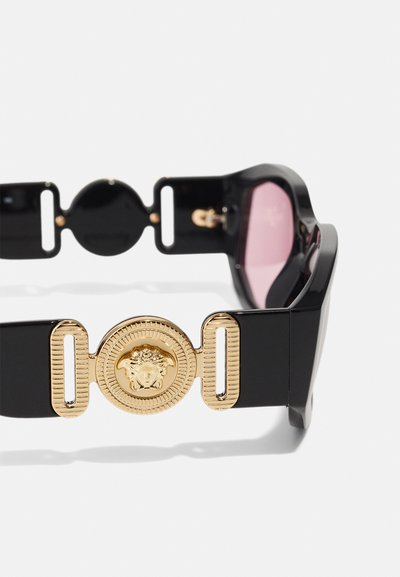Sorte solbriller med pink linser, med et fremtrædende guld cirkulært emblem med tekstureret kant og distinkte hardware detaljer.