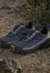 Scarpe da trail running GORE-TEX in blu navy e grigio. Presentano una tomaia texturizzata, dettagli riflettenti e una suola imbottita per aderenza e stabilità.