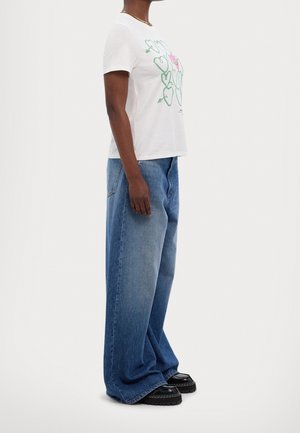 Woman wearing white T-shirt with graphic hearts, baggy blue jeans, and black platform shoes standing sideways on white background.