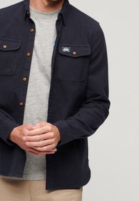Camicia blu navy con due tasche sul petto, bottoni marroni a contrasto e una piccola toppa con logo. Indossata sopra una maglietta grigia.