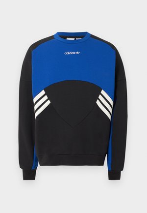Fiatal férfi kék-fekete Adidas pulóverben, fehér csíkokkal, egy egyszerű világos háttér előtt áll.