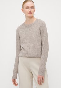 Maglione beige lavorato a maglia con maniche lunghe a raglan e collo rotondo a costine. Indossato con pantaloni chiari e larghi. Texture liscia e morbida.