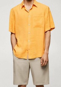 Uomo che indossa una camicia arancione a maniche corte con bottoni e tasca sul petto, insieme a pantaloni corti beige fino al ginocchio, in piedi contro uno sfondo neutro.