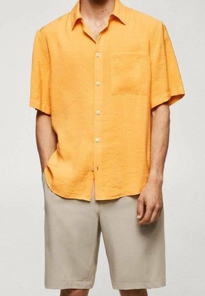 Uomo che indossa una camicia arancione a maniche corte con bottoni e tasca sul petto, insieme a pantaloni corti beige fino al ginocchio, in piedi contro uno sfondo neutro.