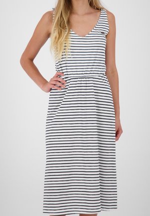 Vrouw draagt een mouwloze, witte midi-jurk met zwarte horizontale strepen, één hand op de heup, tegen een effen witte achtergrond.