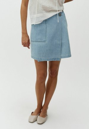 Woman wearing light blue denim wrap skirt with front pocket, white sleeveless blouse, and beige slip-on flat shoes, standing on white background.