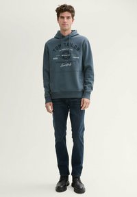 Tume sinine kapuutsiga dressipluus, millel on jaanalinnukotimehhanism, reguleeritav nöör ja trükised. Kombineeritud tumedate teksade ja mustade kingadega. Lihtne, juhuslik stiil.