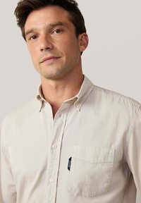 Camicetta button-up beige chiaro con colletto, dotata di una tasca sul lato sinistro del petto e una piccola etichetta blu. Tessuto morbido con una texture liscia.