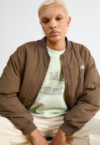 Jaqueta bomber castanha com punhos e colarinho canelados, apresentando um fecho de correr e o logo da New Balance, usada sobre um sweatshirt verde menta com texto.
