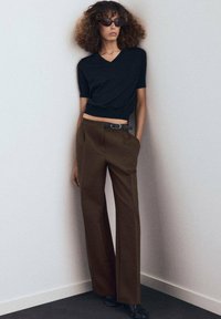 Camisa negra de cuello en V corta combinada con pantalones de pierna ancha marrones de talle alto, acentuada por un cinturón delgado y gafas de sol oscuras.