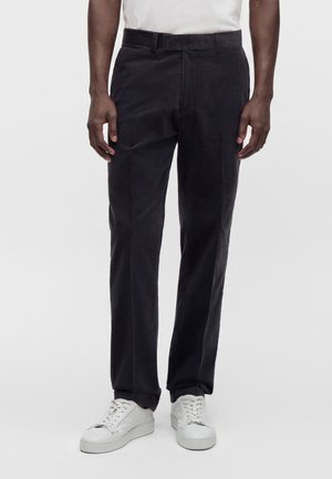 Homme portant un pantalon en velours côtelé noir et des baskets blanches, debout devant un fond clair uni.
