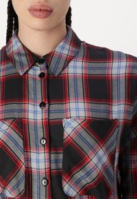 Camicia a quadri rossa e nera con colletto button-down, due tasche frontali e accenti blu a contrasto nel motivo a quadri. Materiale in cotone.