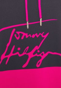 Sweat à capuche noir et rose avec cordons roses et logo "Tommy Hilfiger" en écriture cursive sur la poitrine.