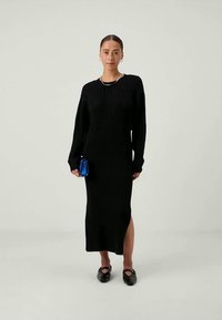 Črna rebrasta pletena obleka z dolgimi rokavi in stranskim razporkom, v kombinaciji z majhno modro torbico in črnimi natikači. Vidna teksturirana tkanina.