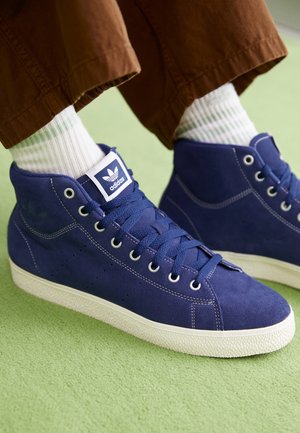 Person iført blå ruskind Adidas high-top sneakers med hvide såler og hvide sokker, stående på en lysegrøn overflade.