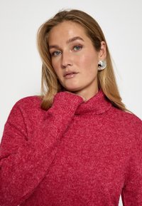 Sarkans turtlenecks džemperis, kas izgatavots no teksturēta trikotāžas auduma, ar garām piedurknēm. Modelis to valkā, pieskaroties kaklam. Redzami sudraba izteiksmīgi auskari.