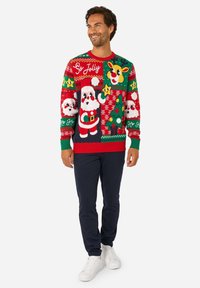 Raštuotas Kalėdinis megztinis raudonos, žalios ir mėlynos spalvų, vaizduojantis Kalėdų Senelį, elnius ir šventinius motyvus. Su ribbintais rankogaliais ir apačia. Dėvimas su tamsiai mėlynomis kelnėmis.