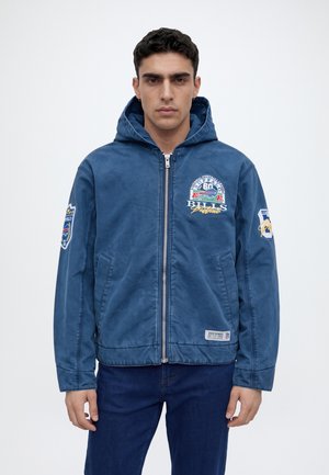 Un homme pose avec une veste bleue à capuche et fermeture éclair, ornée d’écussons de style football, et un jean foncé sur un fond blanc uni.