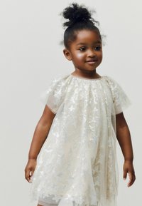 Robe blanche à manches courtes avec un voile léger et transparent présentant des motifs d'étoiles argentées. Texture douce et design ample et fluide.
