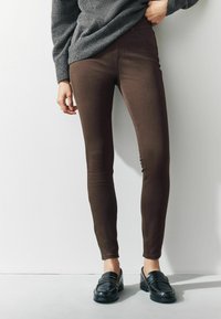 Jeans aderenti marroni con una texture liscia, caratterizzati da una vita media e dettagli di cucitura discreti, abbinati a mocassini neri.