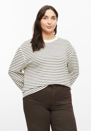 Vrouw met lang donker haar, die een wit-zwart gestreepte trui en donkerbruine broek draagt, staande tegen een effen witte achtergrond.