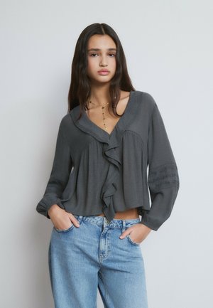 Jeune femme aux longs cheveux noirs portant un chemisier gris foncé à volants et un jean bleu clair, debout les mains dans les poches.