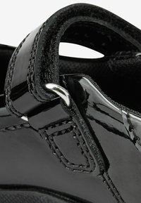 Schwarzer Lacklederschuh mit einem gebogenen Riemen, metallischen Details und sichtbaren Nähten. Texturierte Innenauskleidung.