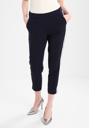 Pantalons bleu marine à coupe détendue, taille élastique, poches latérales et rayures blanches contrastantes sur les côtés. Assortis à des talons argentés.