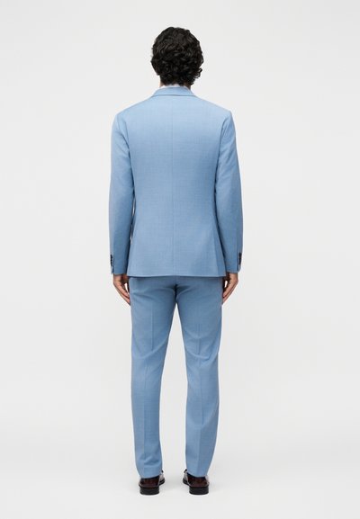 Άνδρας που φαίνεται από πίσω, φοράει ανοιχτό μπλε κοστούμι με ταιριαστό παντελόνι και σκούρα καφέ επίσημα παπούτσια, σε απλό λευκό φόντο.