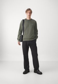 Felpa verde in cotone con collo a giro, pantaloni neri e una borsa a spalla nera. Accenti notevoli includono un piccolo logo sulla felpa.