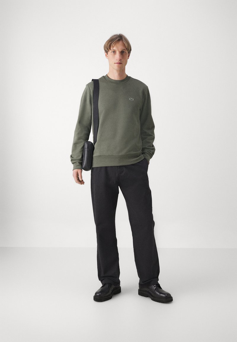 Groene katoenen sweatshirt met ronde hals, zwarte broek en een zwarte schoudertas. Opvallende accenten zijn een klein logo op het sweatshirt.
