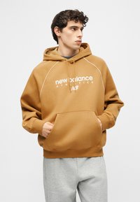 Νέος άνδρας με μπεζ φούτερ New Balance με τα χέρια στην μπροστινή τσέπη, κοιτάζοντας δεξιά, φοράει ανοιχτό γκρι παντελόνι μπροστά σε απλό λευκό φόντο.