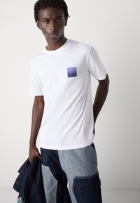 White cotton t-shirt featuring a square blue logo on the chest. The model pairs it with two-tone denim trousers and holds a jacket.