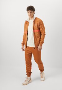 Oranžni trenirka iz gladkega blaga, ki ima bel ovratnik, rožnate črte na prsih in oprijete hlače. Bele superge dopolnjujejo izgled.