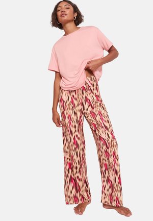 Vrouw poseert op blote voeten met een losse roze T-shirt en wijde broek met roze en zwarte luipaardprint.