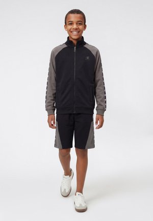 Chłopiec idący do przodu, uśmiechający się, ubrany w czarno-szarą kurtkę na zamek z logo na rękawach, pasujące szorty i białe tenisówki.