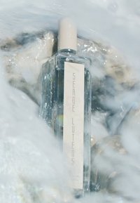 Klarer Glasparfümflacon mit einem beigen Deckel, der ein weißes Etikett mit erhabenem Text trägt, umgeben von Wasser und glatten Steinen.