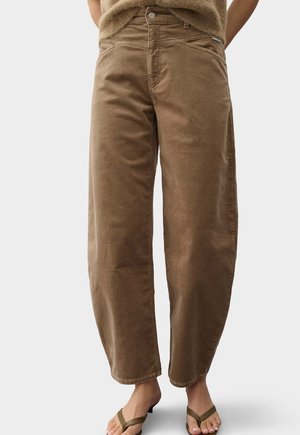 Braune, hoch taillierte, weit geschnittene Hosen, kombiniert mit grünen Flip-Flop-Sandalen, dargestellt von der Taille bis zu den Füßen vor neutralem Hintergrund.