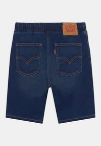 Tummansiniset farkkushortsit rennolla istuvuudella, joissa on kaksi takataskua, keltaista tikkausta ja ruskea nahkainen logopainatus vyötäröllä.